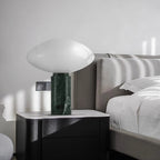 Clérachic table lamp lit on a bedside cabinet next to a grey upholstered bed, its pale marble base beside a wall socket.