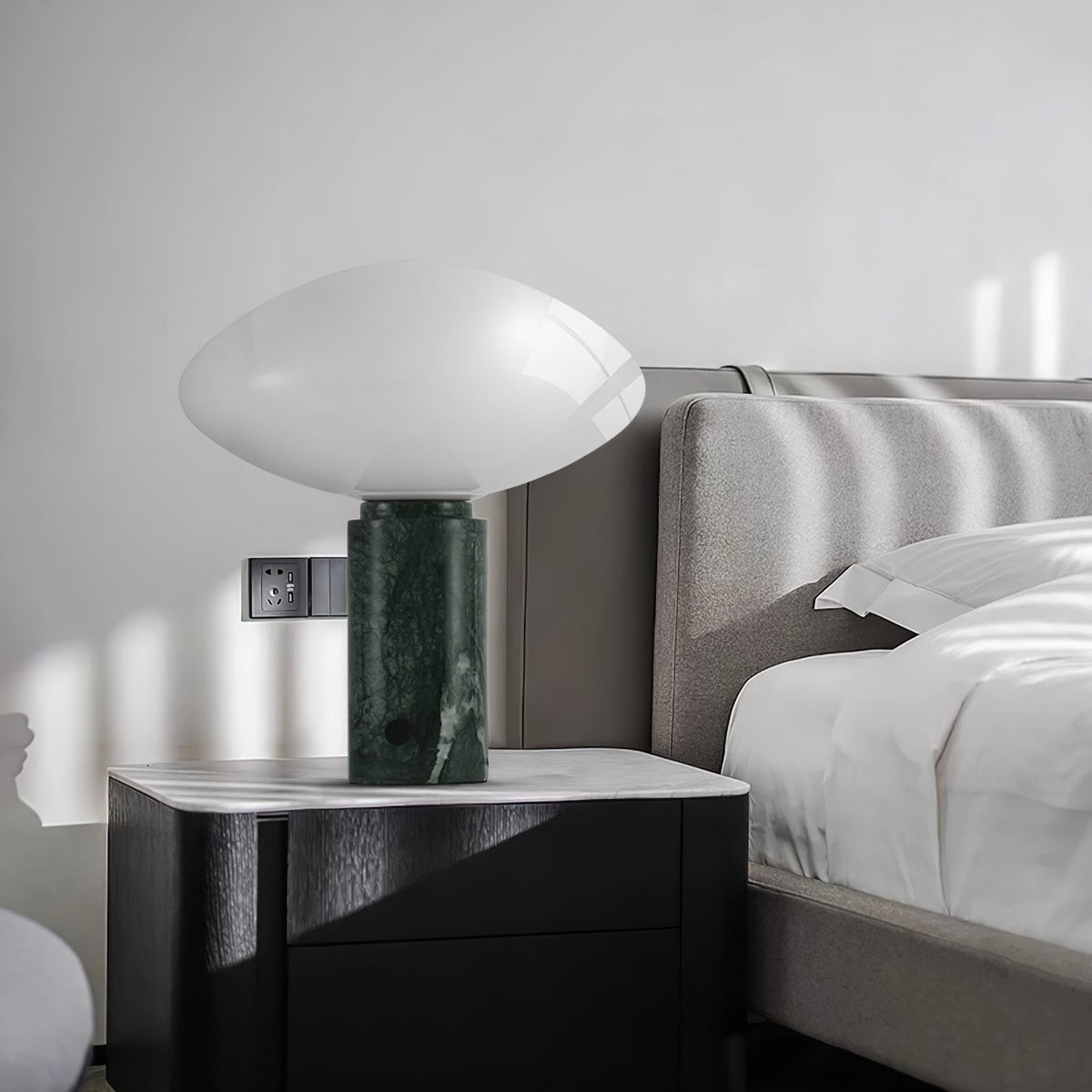 Clérachic table lamp lit on a bedside cabinet next to a grey upholstered bed, its pale marble base beside a wall socket.