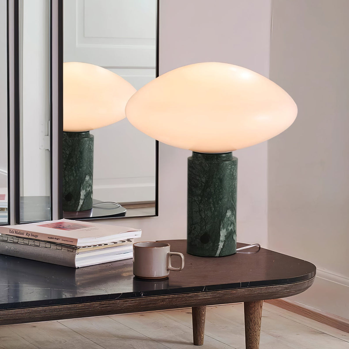 Clérachic table lamp reflected in a leaning mirror on a dark wood table, with a mug and open books beside it.