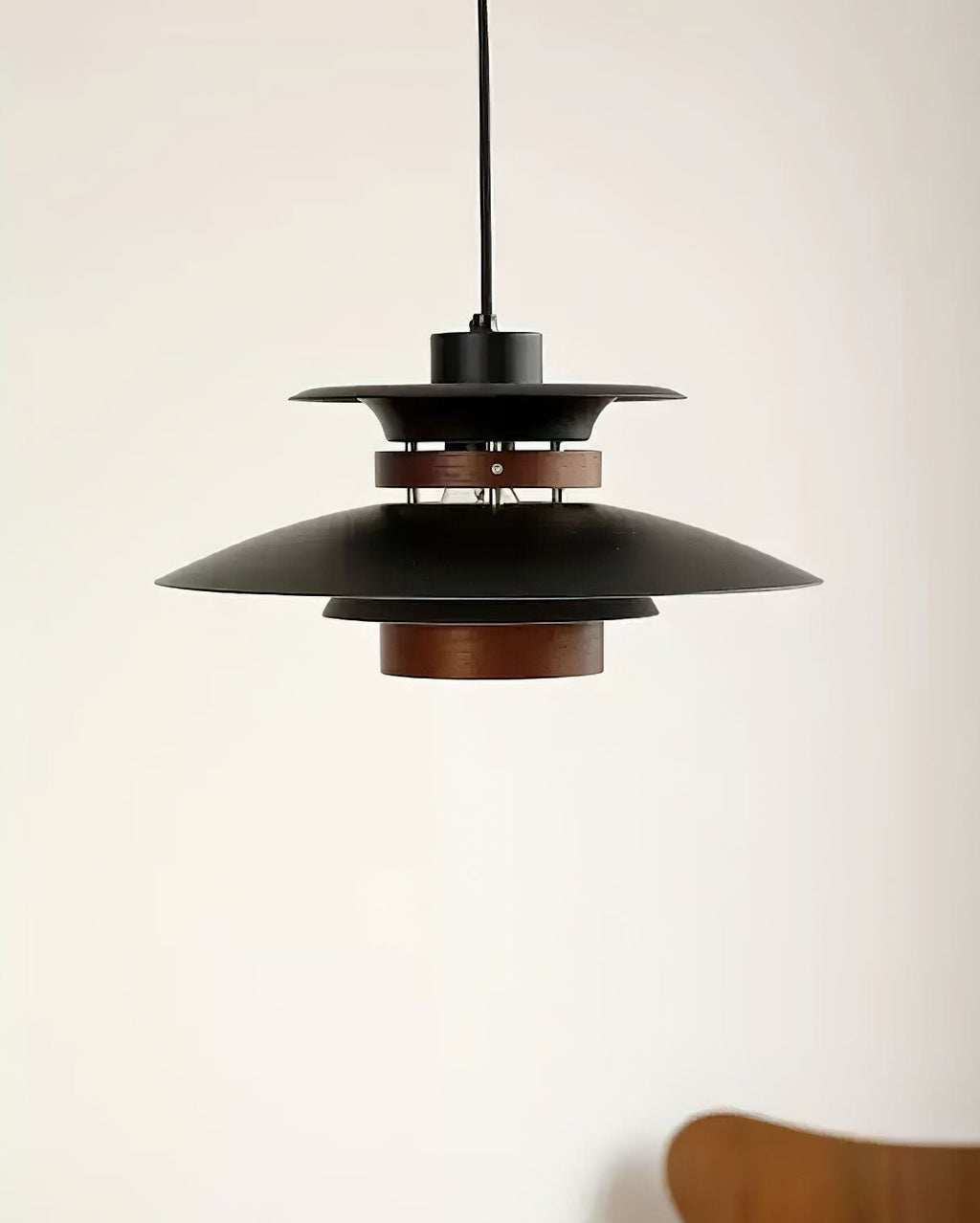 A black Clérachic pendant hangs against a plain wall beside a wooden chair.