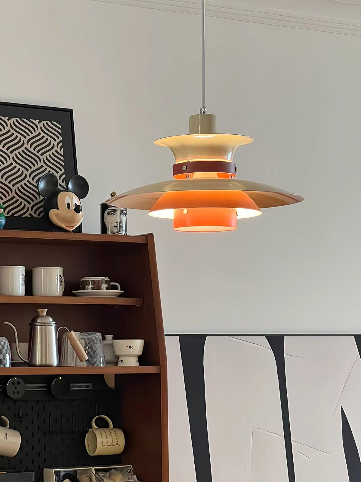 A lit beige Clérachic pendant hangs over open kitchen shelving with mugs and a kettle.