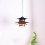 A lit black Clérachic pendant hangs in a plain room with a leafy plant nearby.