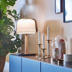 Clérachic lamp lit on a blue sideboard beside pillar candles and a potted plant, glass shade glowing warm.