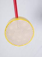 Close-up of a single cane drum shade on a Clérachic chandelier's red arm, its perforated rattan weave lit from within against a yellow brass rim.