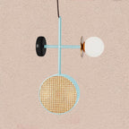 Clérachic pendant with a black-and-teal cross-rod, a cane drum shade below and a frosted glass globe to the side, set against a textured pink wall.