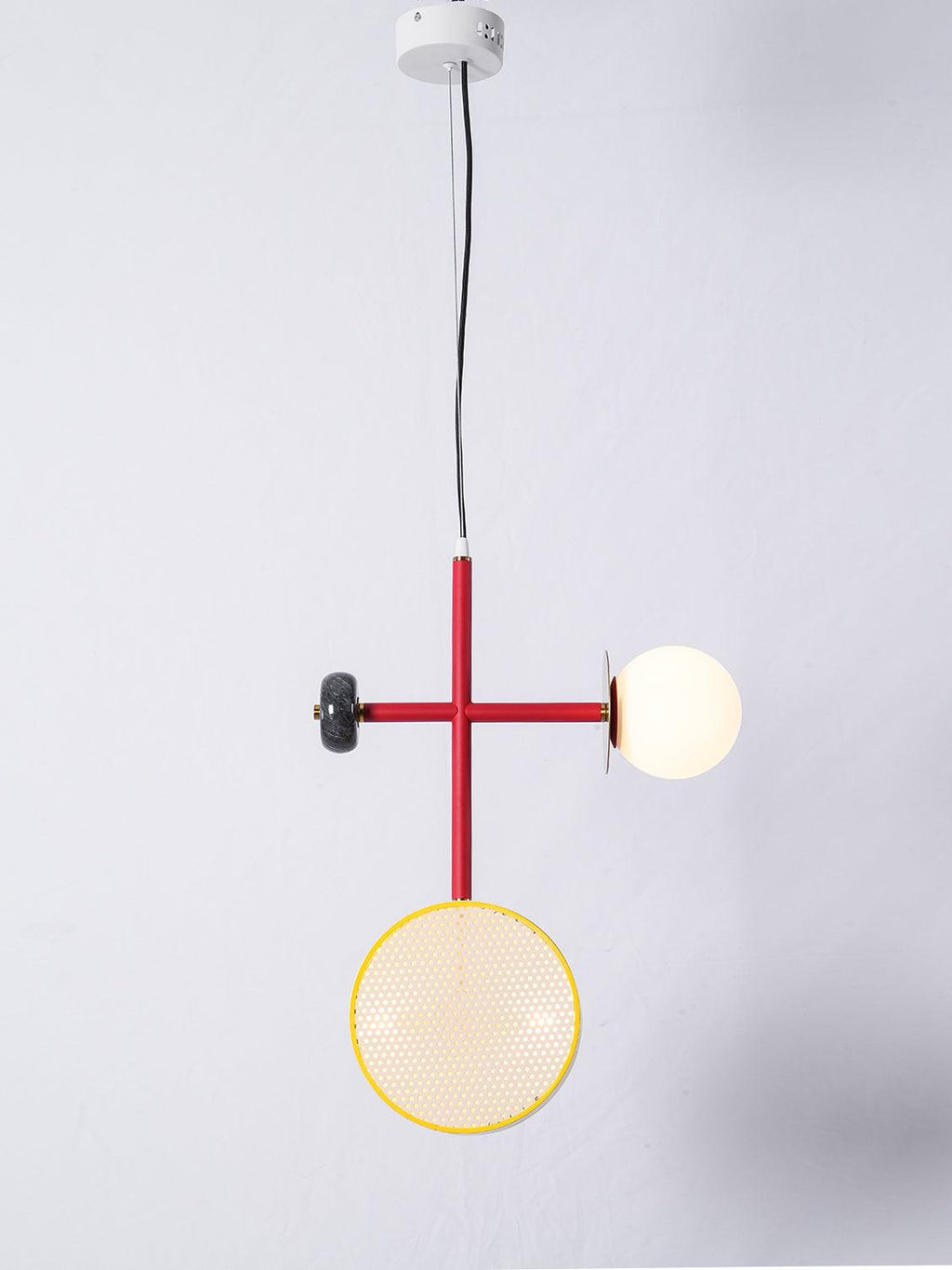 Small two-armed Clérachic pendant with a red cross-rod, a yellow-rimmed cane drum below and an opal glass globe to the side, on a plain grey wall.