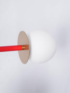 Extreme close-up of a Clérachic pendant's red rod meeting a brass disc and frosted glass globe against a plain white wall.