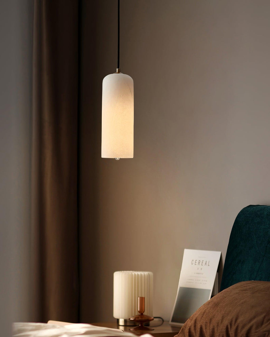 The Zashayra pendant lit in a dim bedroom corner beside a ribbed table lamp and a magazine.