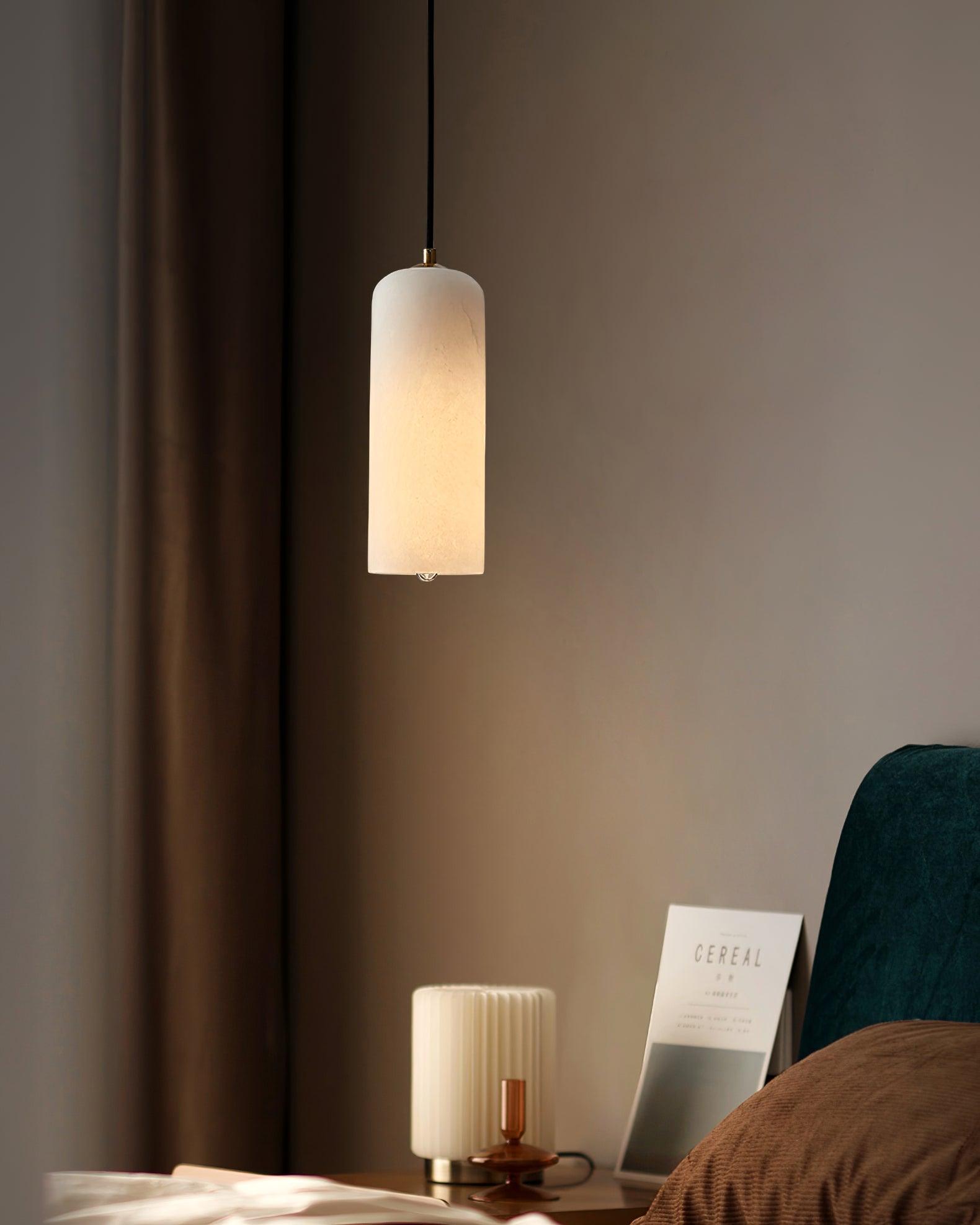The Zashayra pendant lit in a dim bedroom corner beside a ribbed table lamp and a magazine.