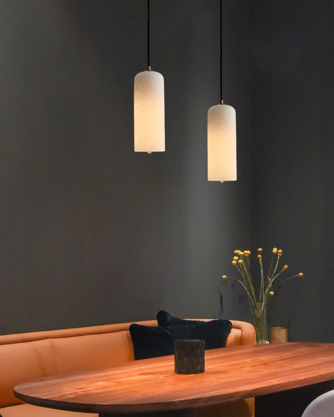 A pair of Zashayra pendants lit above an oval wooden dining table against a dark wall.