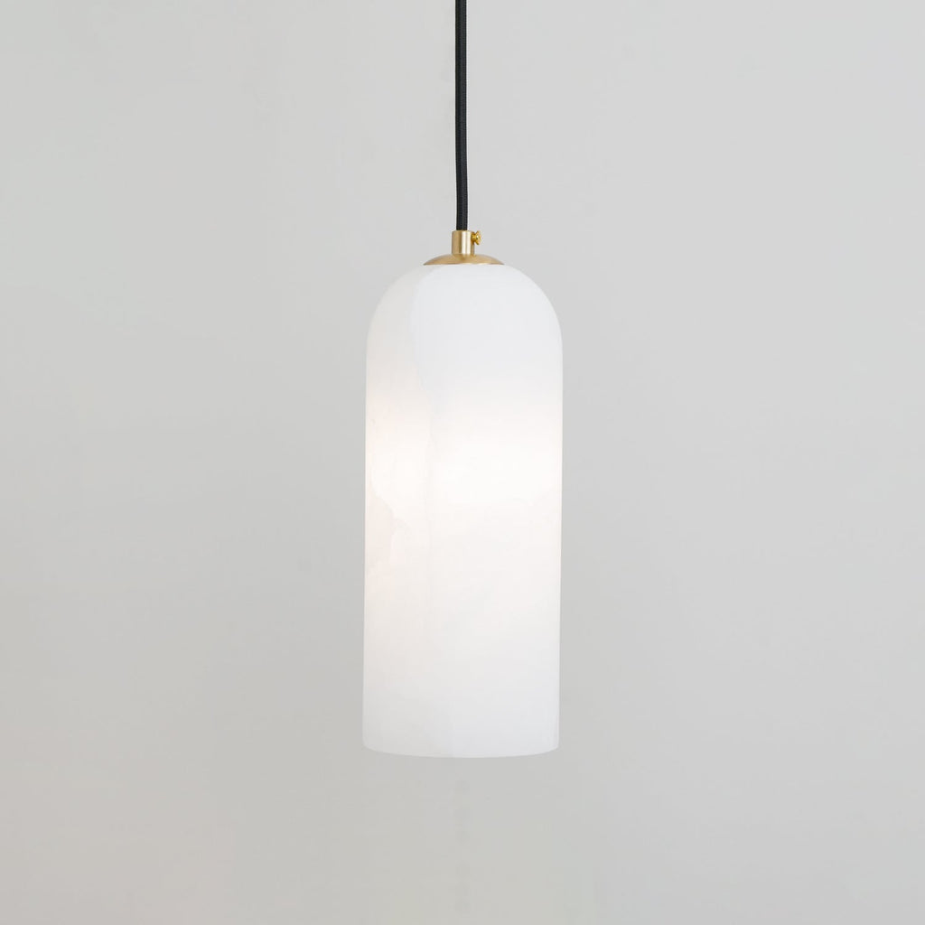 Hecate pendant lit softly against a pale grey wall, brass cap visible where the black cord fits.