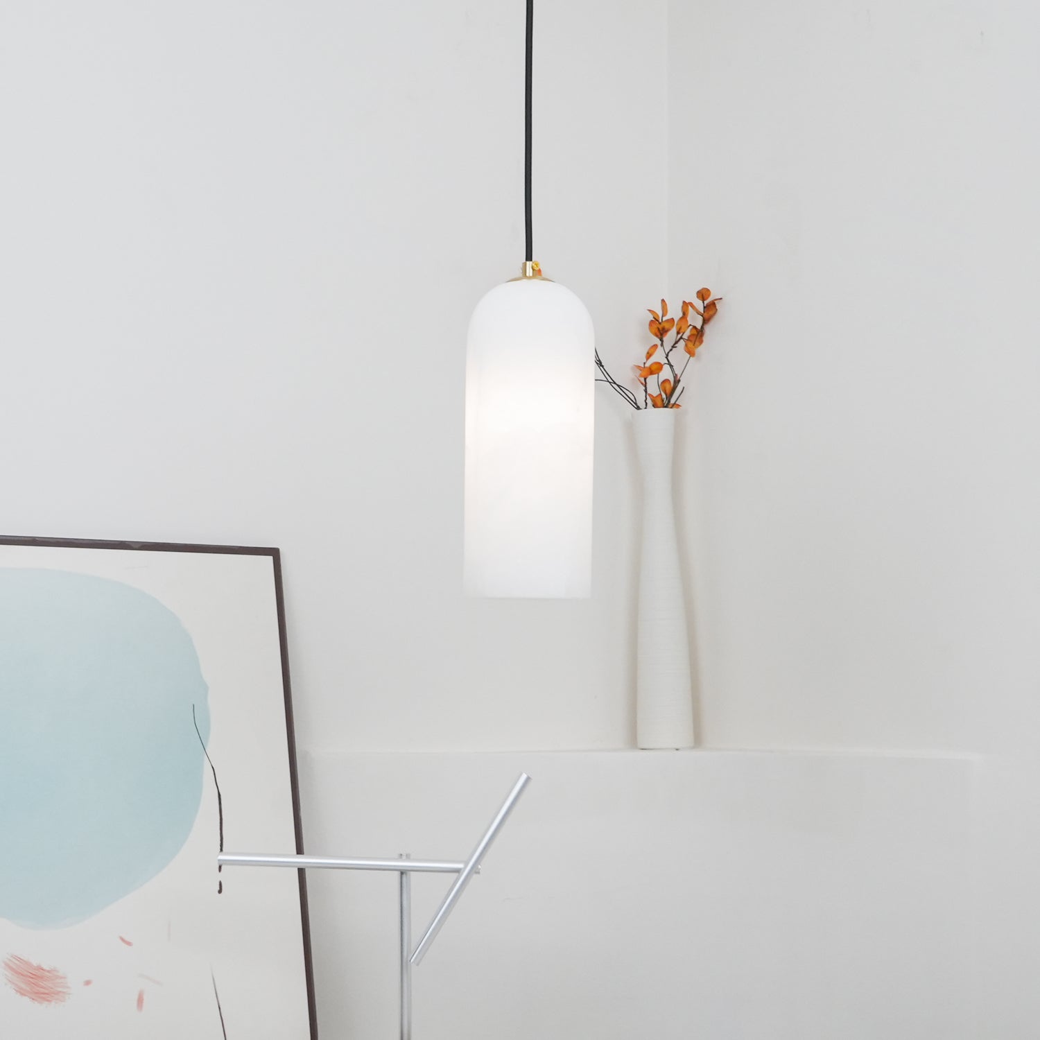 Zashayra pendant lit in a bright white corner above a ribbed vase and a leaning framed print.