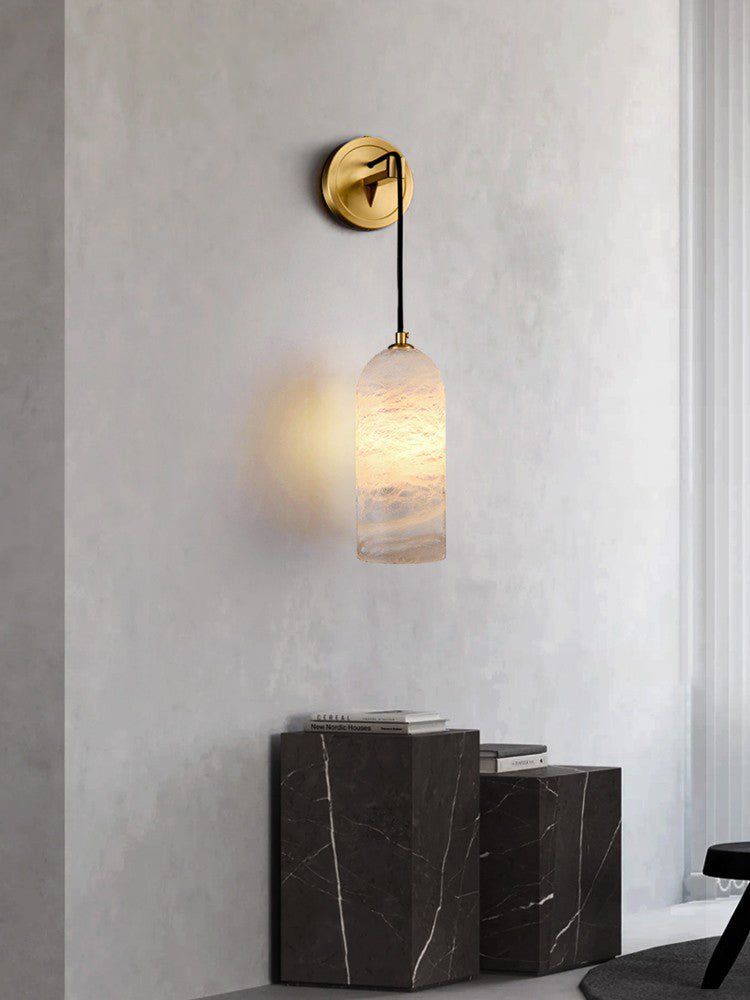 Vaelara sconce glowing on a concrete-toned wall above two black marble plinths stacked with books