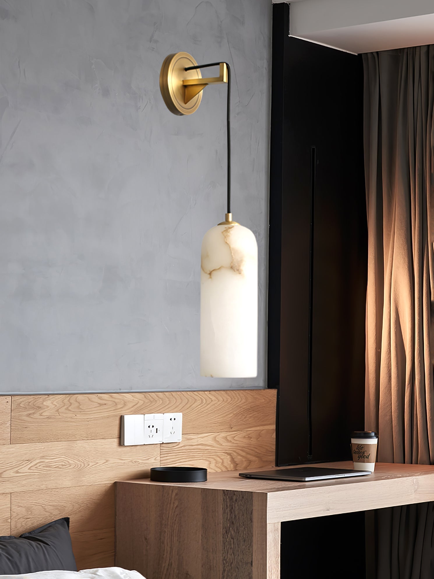 Vaelara sconce mounted on a grey plaster wall above an oak bedside desk with a laptop and coffee cup