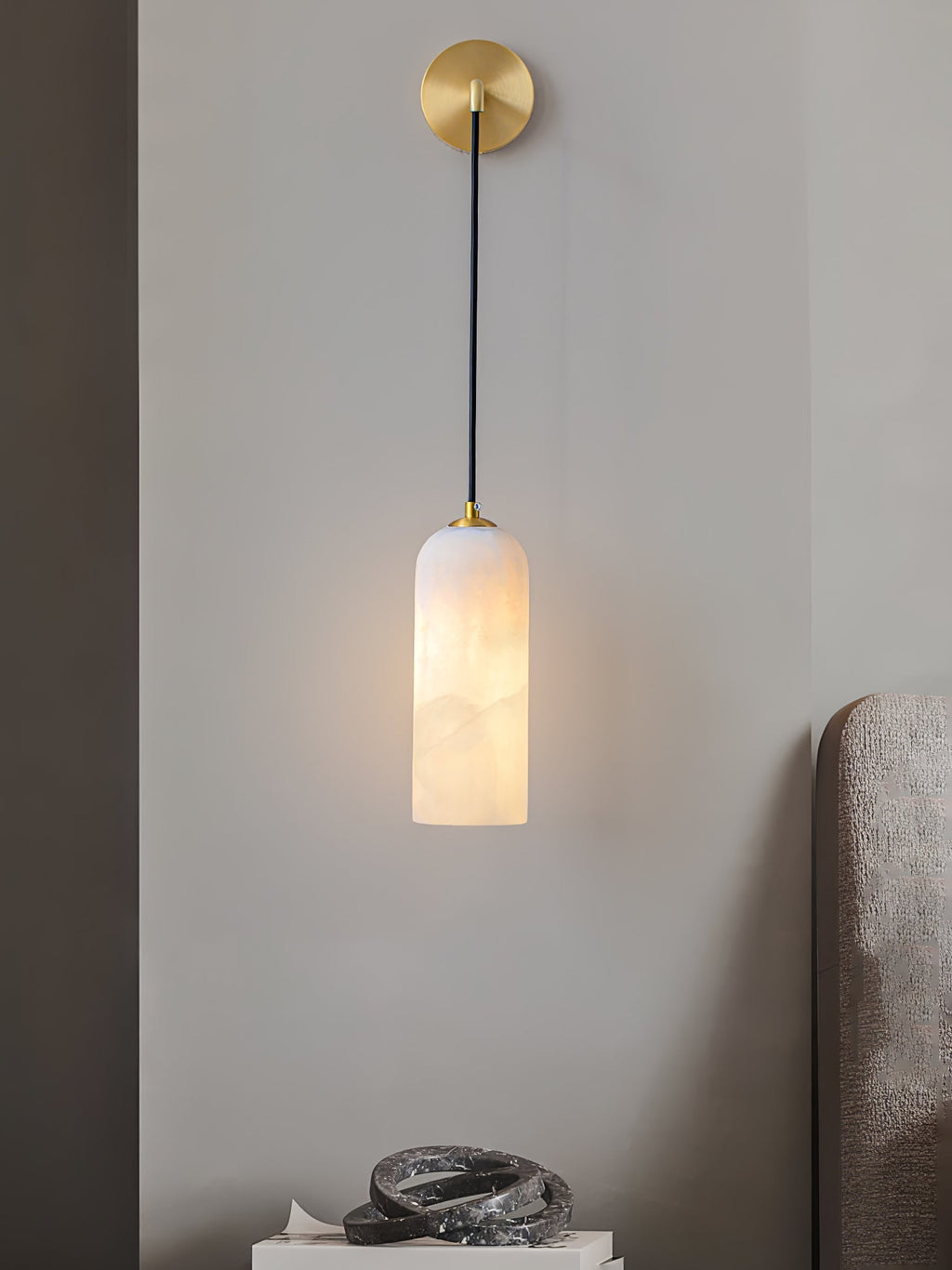 Vaelara alabaster sconce lit beside a bed, its glow washing the grey wall above a marble sculpture