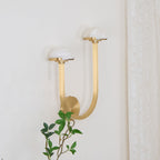 Brass Vitalira Double Alabaster Wall Light lit on a cream wall beside a lace curtain and leafy branch.
