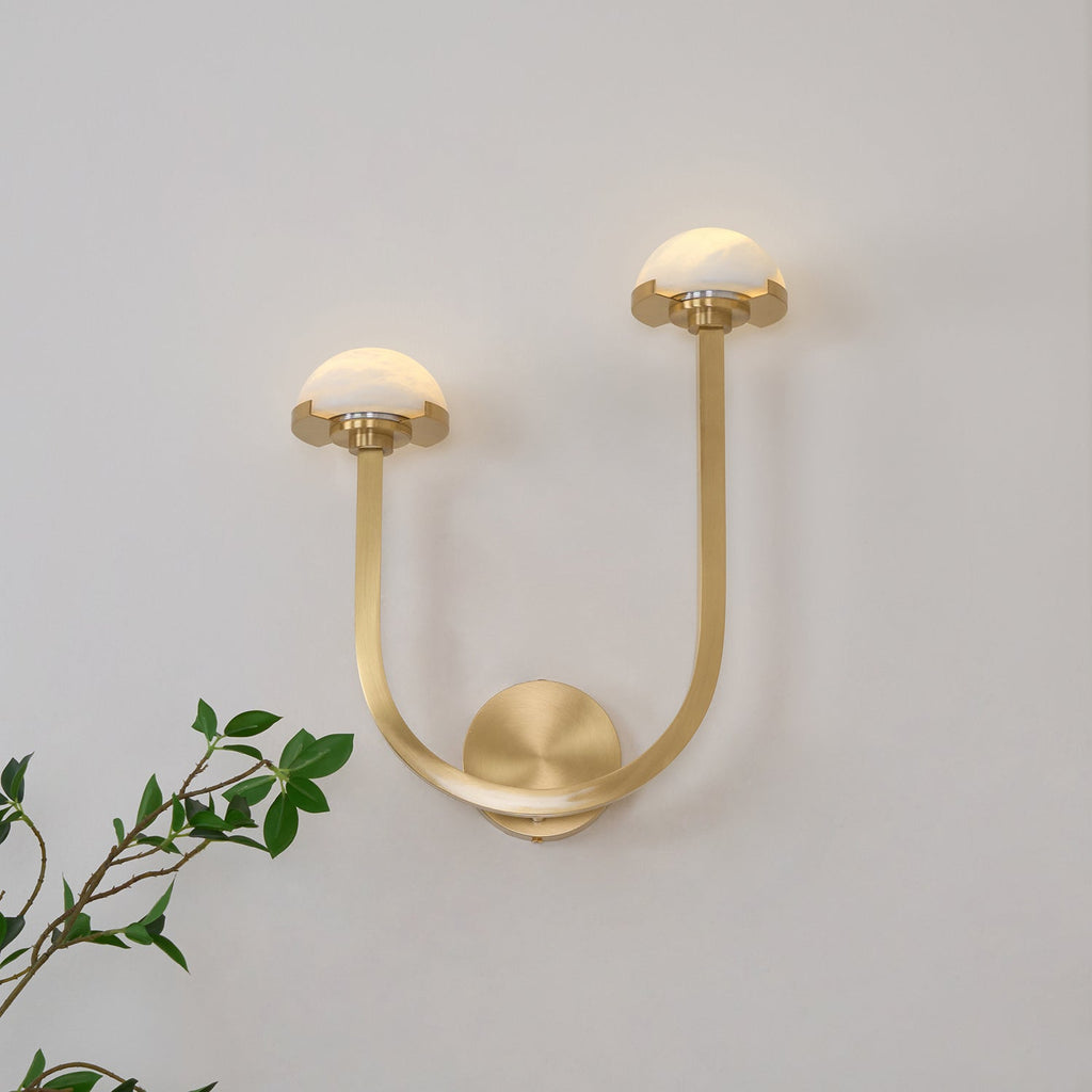 Satin brass Vitalira Double Alabaster Wall Light lit on a white wall with a green branch in frame.