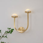 Satin brass Vitalira Double Alabaster Wall Light lit on a white wall with a green branch in frame.
