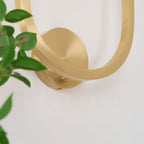 Detail of the round brushed brass wall plate and the curved arm crossing it on the Vitalira wall light.