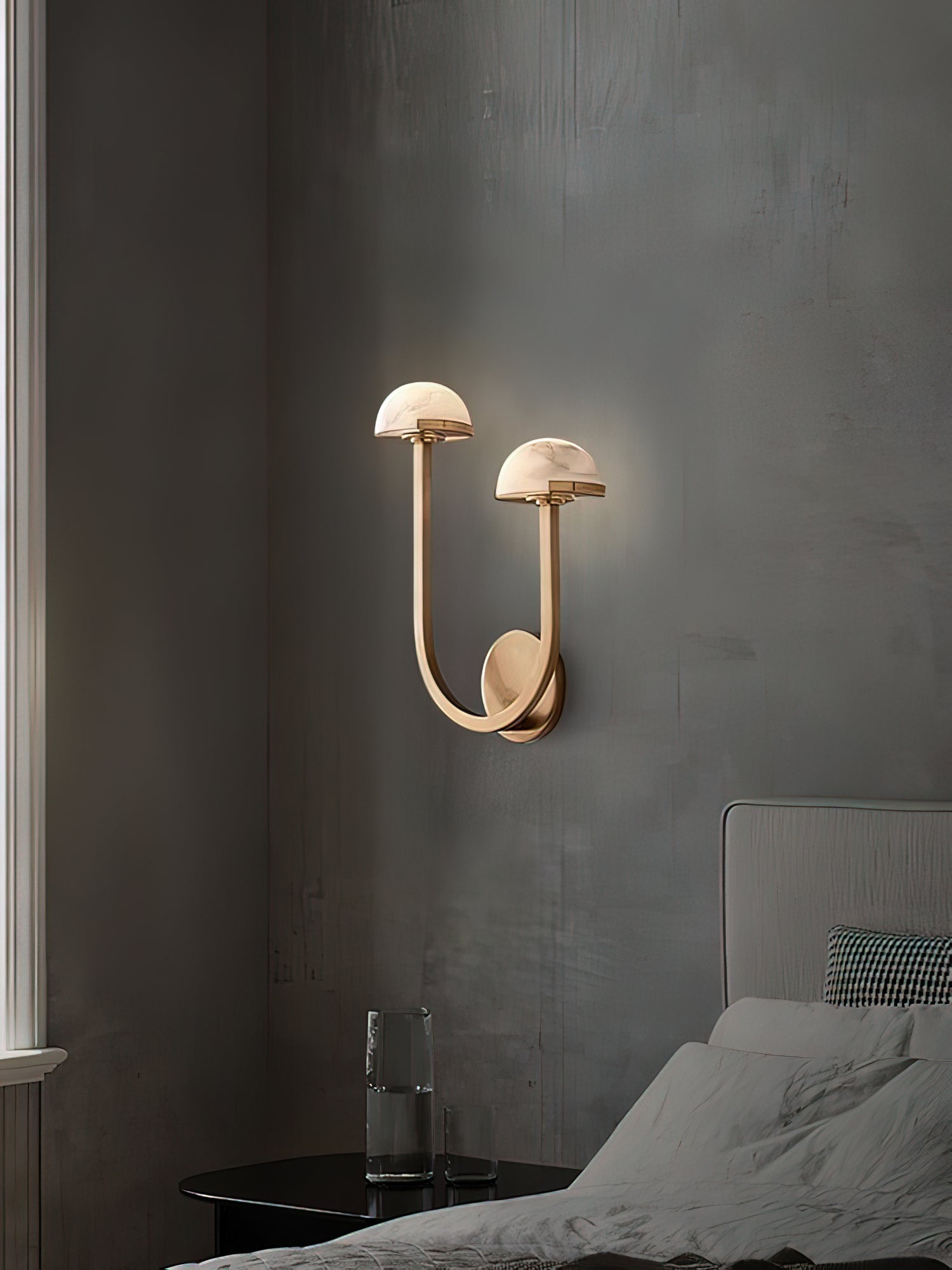 Brass Vitalira Double Alabaster Wall Light lit in a grey bedroom corner beside a nightstand and glass.