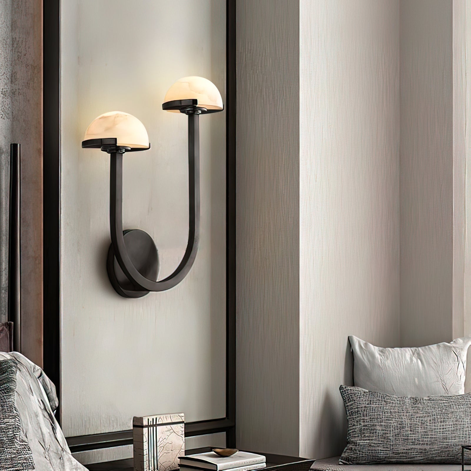 Black Vitalira Double Alabaster Wall Light lit on a pale panelled wall between two textured cushions.