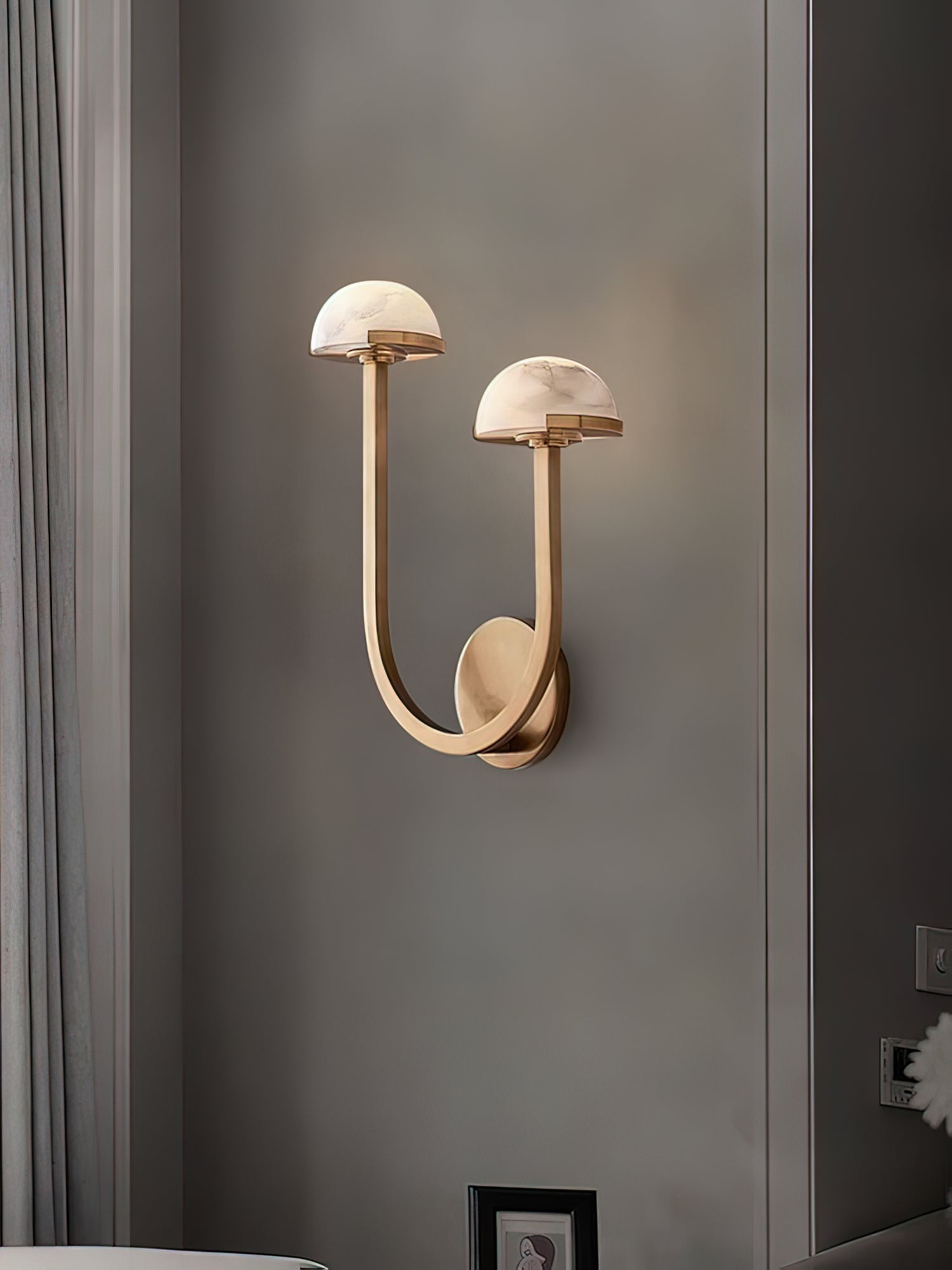 Brass Vitalira Double Alabaster Wall Light lit on a grey wall, framed picture and curtain nearby.