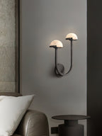 Black Vitalira Double Alabaster Wall Light lit above a bedside table in a dim, grey-walled bedroom.
