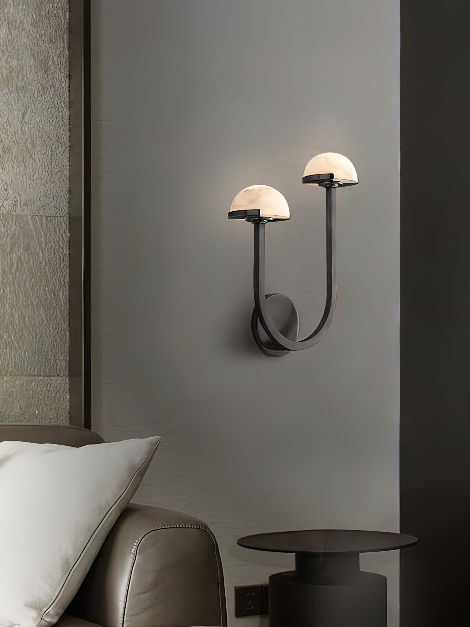 Black Vitalira Double Alabaster Wall Light lit above a bedside table in a dim, grey-walled bedroom.