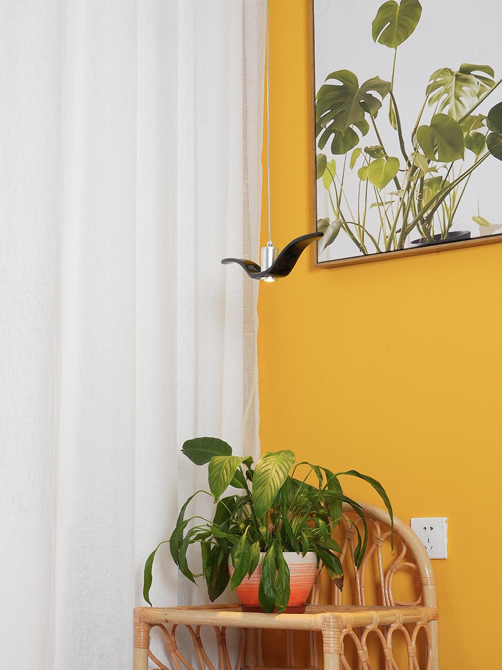 A black bird-shaped pendant from this Clérachic light hangs near a mustard-yellow wall with potted plants and a sheer curtain.