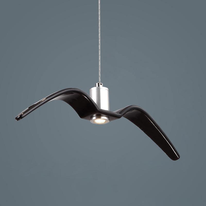 A black bird-shaped pendant from this Clérachic light hangs with its wings swept back, on a dark blue-grey backdrop.