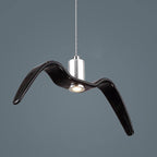 A black bird-shaped pendant from this Clérachic light hangs at an angle with its LED lit beneath one wing, on a dark blue-grey backdrop.