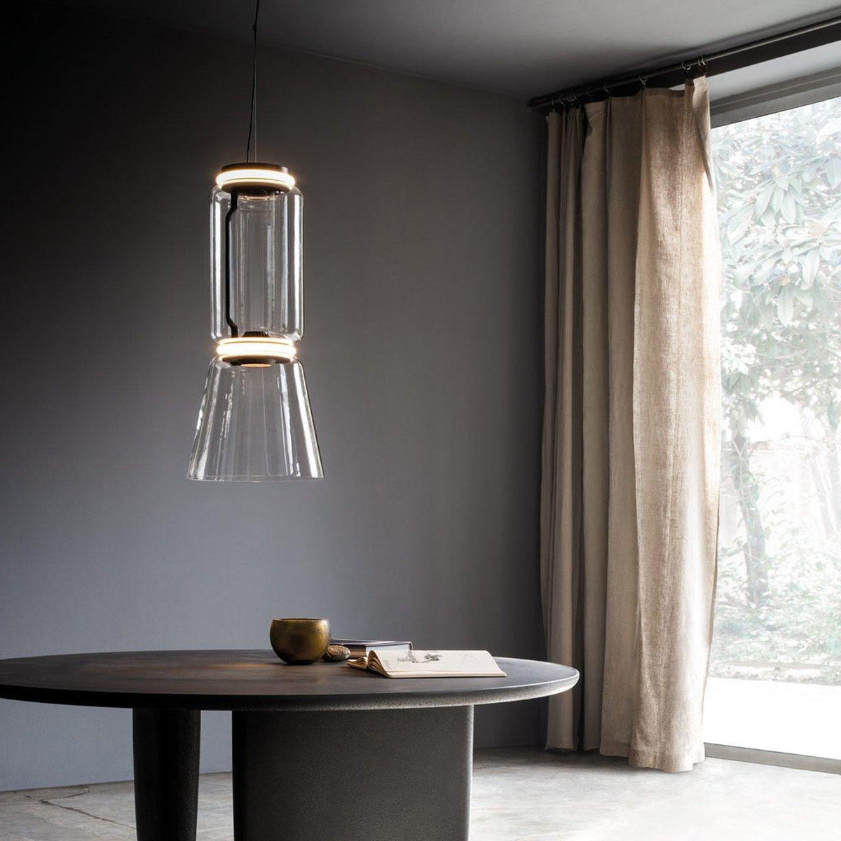 A cone-shaped clear-glass shade on this Clérachic pendant casts warm light over a round table beside a curtained window.