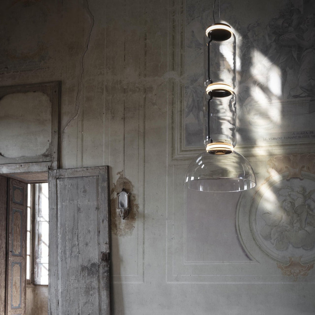 Two Clérachic pendants, a cylinder stack above a dome shade, catch sunlight on a weathered plaster wall.