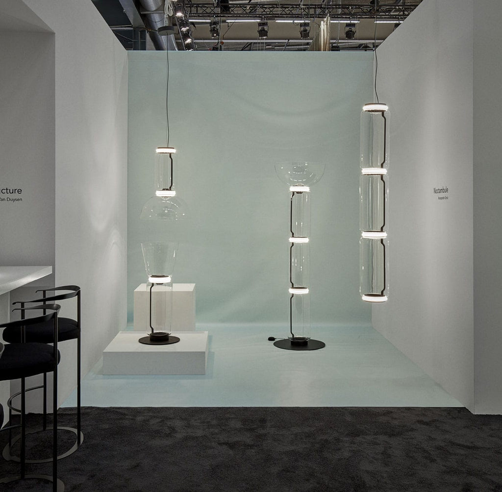 In a pale blue exhibition space, this Clérachic pendant's cylinder stack hangs beside a bell-jar floor fixture.