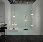 In a pale blue exhibition space, this Clérachic pendant's cylinder stack hangs beside a bell-jar floor fixture.
