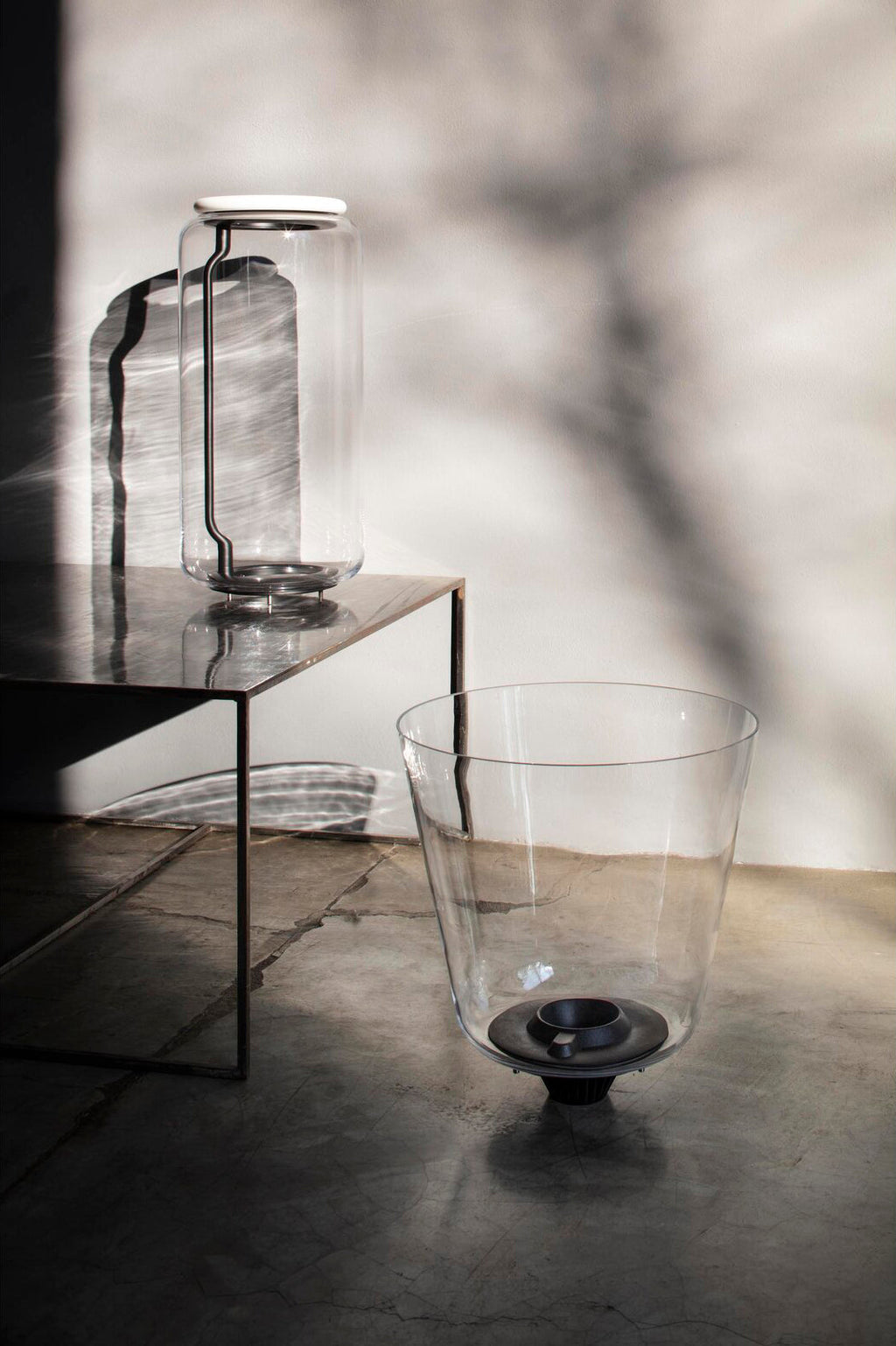 This Clérachic pendant's glass shade sits on a low table beside a wide glass vessel, with branch shadows behind.