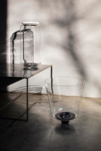 This Clérachic pendant's glass shade sits on a low table beside a wide glass vessel, with branch shadows behind.
