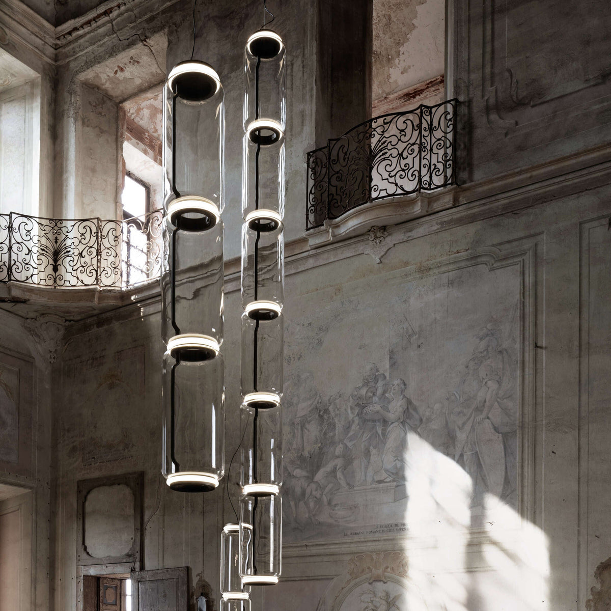 A row of glowing cylinder stacks from this Clérachic pendant hangs between wrought-iron balconies in a frescoed hall.