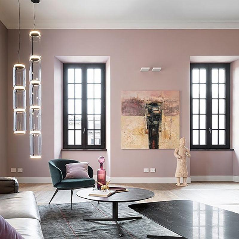A stack of glowing cylinders from this Clérachic pendant hangs near tall windows in a mauve sitting room, beside framed art.