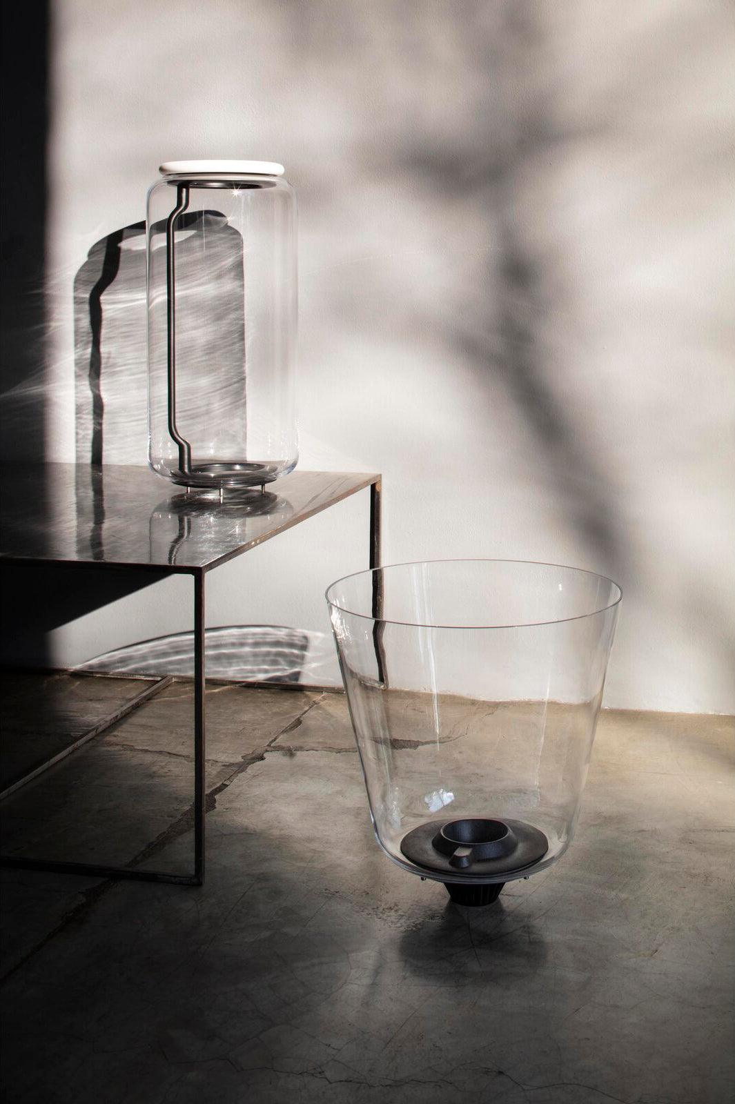 This Clérachic pendant's glass shade sits beside a glass vessel on a low table, with branch shadows on the wall.