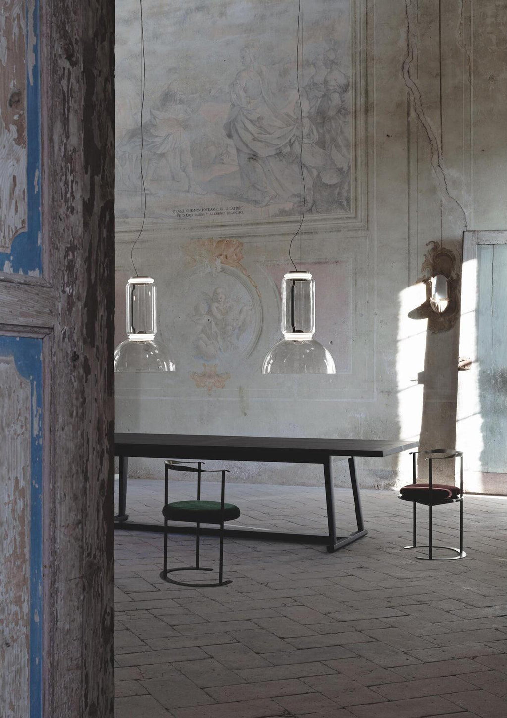 Two single-cylinder Clérachic pendants hang unlit in a weathered outdoor courtyard beside a wire chair and low stool.