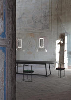 Two single-cylinder Clérachic pendants hang unlit in a weathered outdoor courtyard beside a wire chair and low stool.