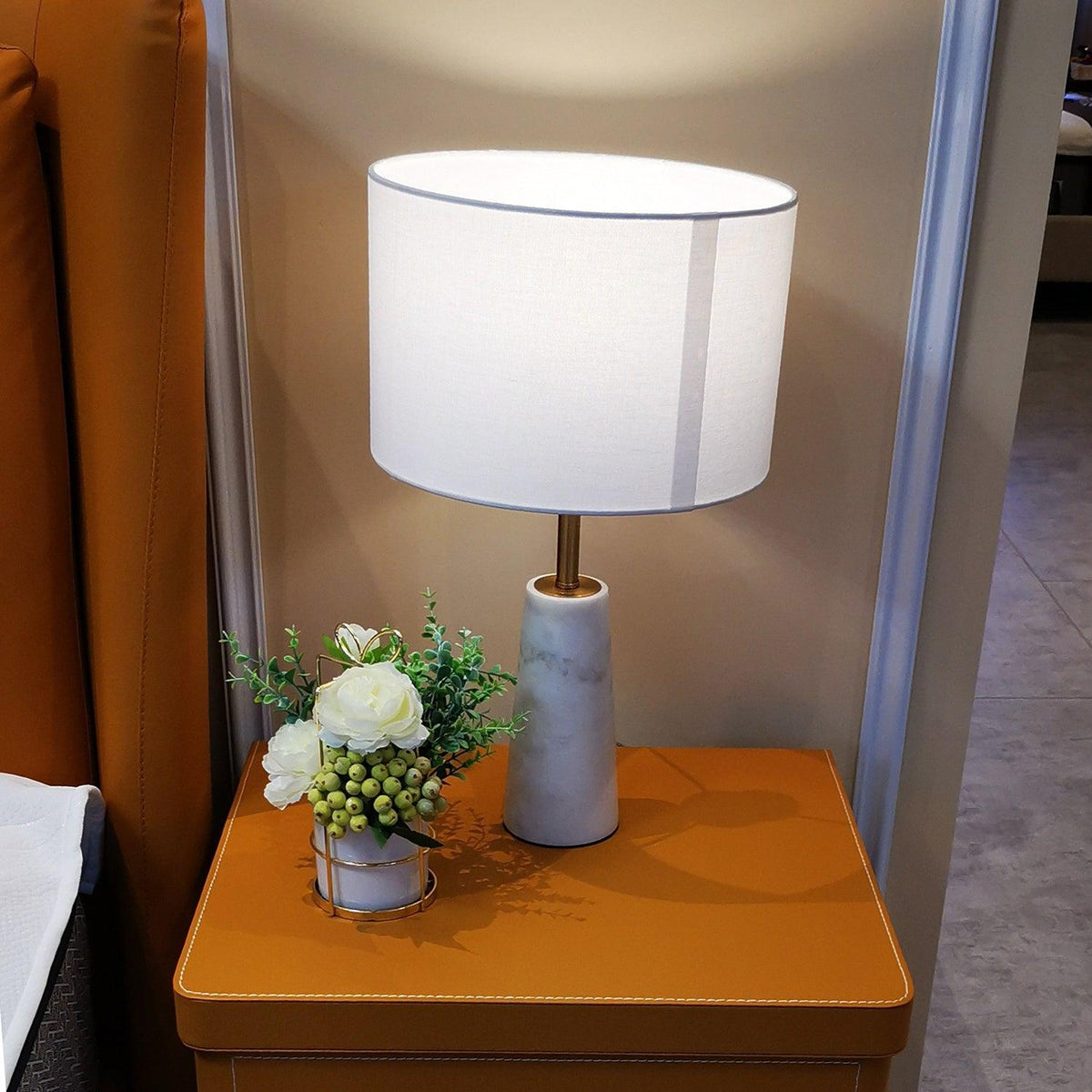Clérachic lamp glowing on a tan leather bedside table next to a small vase of white flowers.