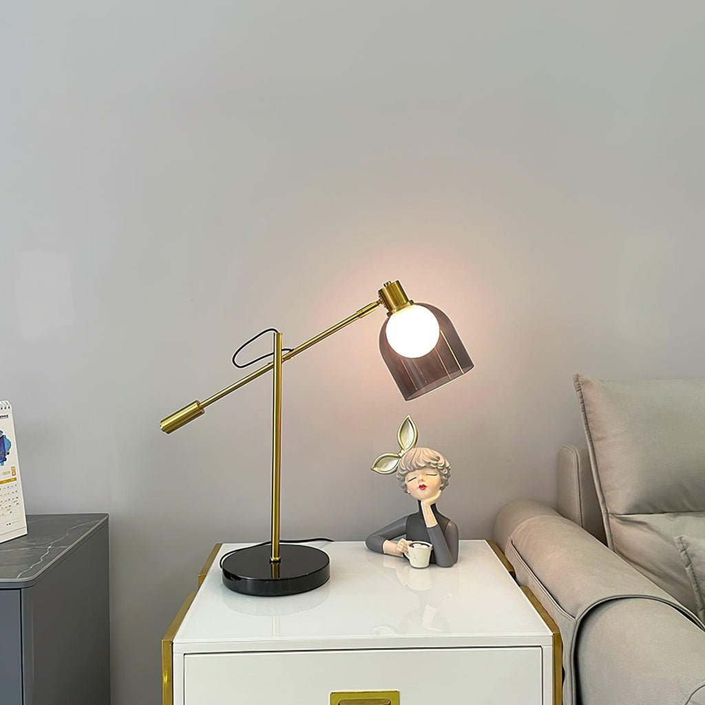 Clérachic brass table lamp lit beside a small figurine on a white bedside cabinet, with a sofa visible behind it.