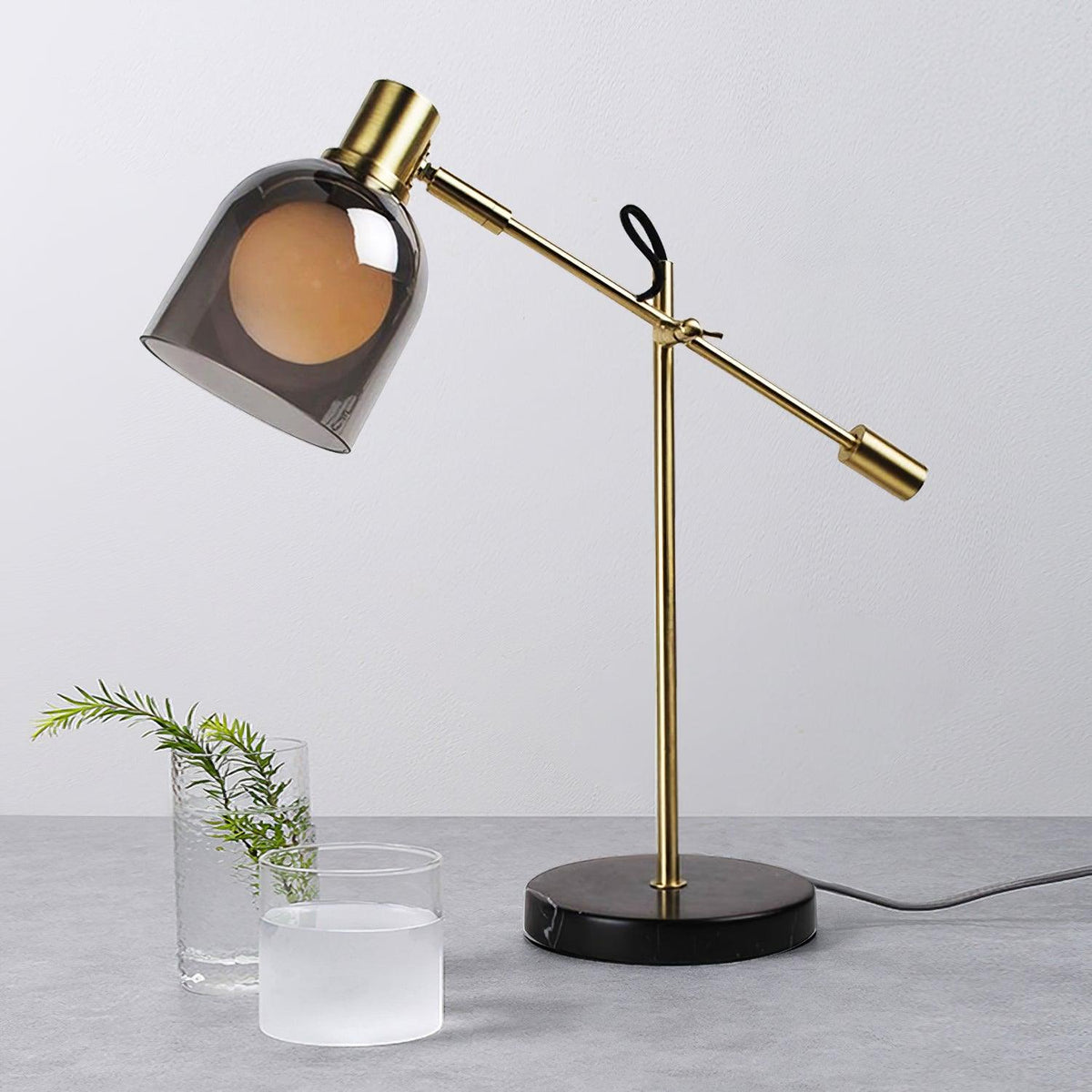 Clérachic brass table lamp beside a glass vase of foliage and a frosted glass votive, styled on a pale grey stone surface.