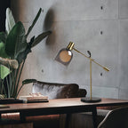 Beside a potted banana-leaf plant, a Clérachic brass table lamp sits on a wood table against a textured concrete wall.