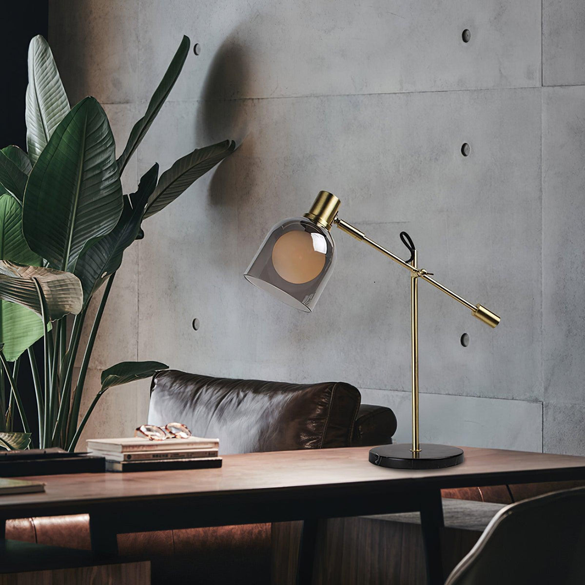 Beside a potted banana-leaf plant, a Clérachic brass table lamp sits on a wood table against a textured concrete wall.