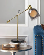 A round mirrored side table stacked with books holds the Clérachic brass table lamp, beside a blue velvet tufted chair.
