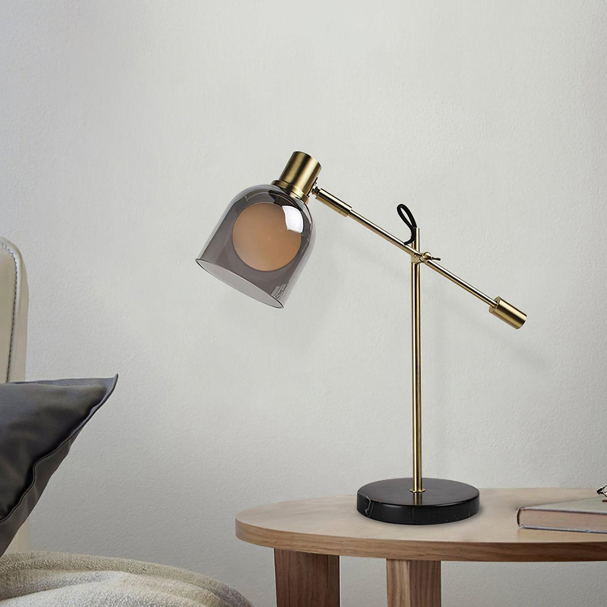 Beside a cushioned sofa and a stack of books, a Clérachic brass table lamp stands on a round wood side table, against a pale wall.