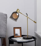 Clérachic brass table lamp on a round black side table beside a framed photo and loose papers, next to a grey sofa.